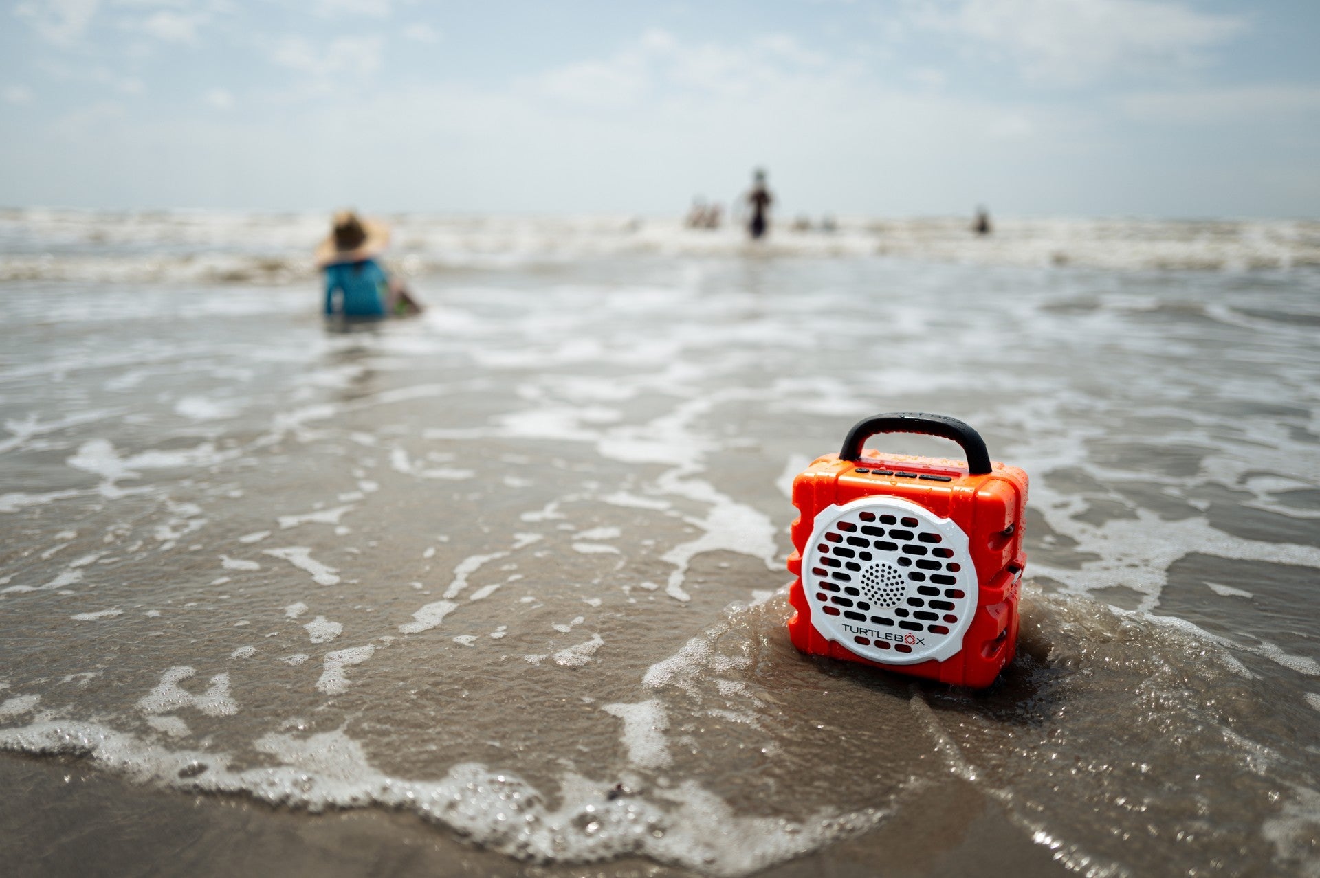 A lifestyle photo of an orange portable radio on a beach with people in the background #base-color_orange