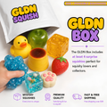 GLDN Squishy GLDN Box surprise toy set shown with a white bag opening, a yellow rubber duck, a green apple-shaped squishy, a translucent yellow cube, a yellow cup, a blue flower-shaped squishy held in a hand, a strawberry-shaped squishy, and a paw-print-shaped squishy, with promotional text on a white background.