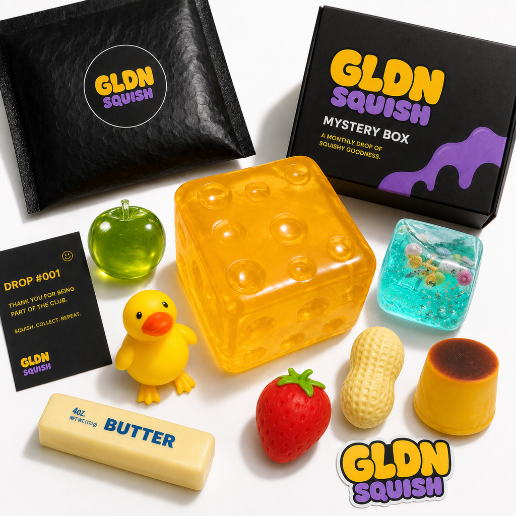 A GLDN Squish Mystery Box set featuring a large translucent yellow cheese-shaped gummy, a small yellow duck figure, a green apple gummy, a blue gel cube, a peanut-shaped gummy, a strawberry gummy, a yellow chocolate gummy, a butter-shaped gummy, and two black branded boxes on a white background.